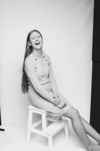 A black and white picture of a long-haired woman laughing on a white stool in front of a white backdrop.