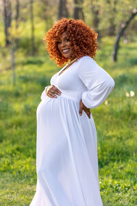 Pregnant woman in a white gown standing with hand on hip in a green field during a Cleveland maternity photography session