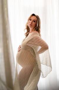 Pregnant woman standing in front of sheer white curtains, softly draped in a translucent gold-toned gown, glowing in natural window light during a fine art maternity session in Waukesha, WI.