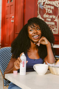 Model at a café table with coffee and Evian facial spray, red door behind