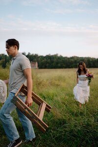 A candid moment captures the scene as a guy carries props after an engagement session in the field, while the girl in a white dress follows him
