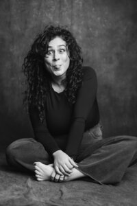 A black and white picture of a woman with dark curly hair sitting cross-legged on a dark gray backdrop. She wears jeans and a black long-sleeve shirt. She wears an amusing expression with her eyes slightly crossed and tongue sticking out of her mouth.