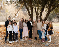 Multigenerational family gathered outdoors beneath large trees with coordinated neutral outfits, joyful candid moment captured by Kaitlyn Dawn Photography, outdoor family portrait specialist.