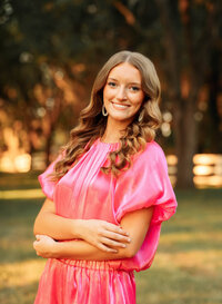 Senior portrait in downtown Magnolia Springs, Alabama — wearing a bright pink outfit, arms crossed.