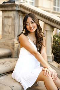 Close-up portrait of a smiling high school senior girl sitting on stone steps in a white dress, senior photographer Kaitlyn Dawn Photography.