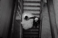 Vue en plongée des mariés se tenant la main dans un escalier métallique, robe volumineuse et complet sombre, moment tendre au Belvédère du Centre des Sciences, Montréal