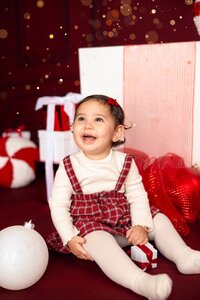 A baby smiles cheerfully. She sits in front of a peppermint pillow and red and white Christmas gifts.