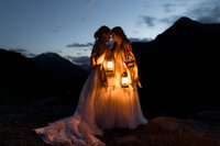 Lesbian couple shown kissing at night holding lanterns with silhouettes of mountains in the background.