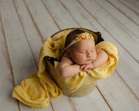 Sleeping newborn baby girl wrapped in a golden yellow swaddle and posed in a wooden bowl prop during a studio newborn photography session.