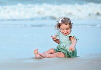 Curly-haired toddler in a mint green dress sitting in shallow ocean water Beach, smiling as she curiously explores the sand during a child portrait session. (Photographed by Nature's Child Photography)