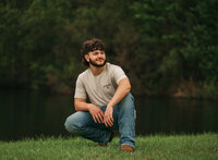 Senior portrait at Circle B Lake in Wilmer, Alabama — crouching in the grass with a pond and
greenery in the background.