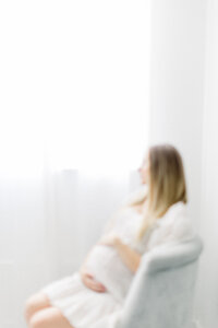 Maternity photo taken in a Dallas photography studio of a pregnant mom sitting on a chair looking outside of the window while she is holding her belly.