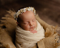 Sleeping newborn baby girl wrapped in a cream swaddle and wearing a floral crown during a studio newborn portrait with newborn photographer Kaitlyn Dawn Photography.