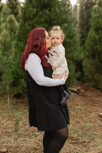 Mother holding her baby in front of evergreen trees, soft natural light family portrait by Kaitlyn Dawn Photography, children’s and family photographer.