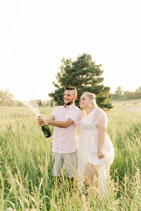 Bride and groom celebrate their engagement session while popping a bottle of champagne in a spring field