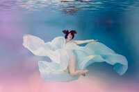 A pregnant mom posing underwater for a maternity portrait session wearing a white bodysuit and long white skirt flowing in the water. By Corrine Studios in Lakeland Florida