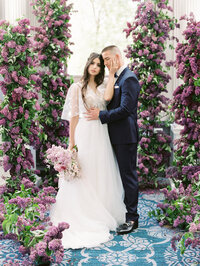 Bride and groom standing in a lush lilac floral ceremony aisle with romantic purple blooms and soft natural light