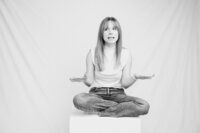 A black and white picture of a blonde woman with straight hair sitting, cross-legged on a white box in front of a white backdrop. She wears a white shirt and jeans. She gestures with her hands out by her sides.
