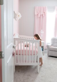 Professional photography by Nature's Child Photography: St. Johns newborn capturing a quiet sibling moment as a little girl gazes into the crib at her baby sister, surrounded by soft pink nursery details in their home.