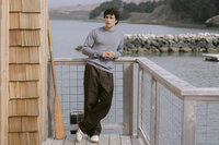 Model in a grey merino crewneck and chocolate brown wide-leg trousers on a dock railing above the bay