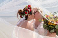 Professional photography by Nature's Child Photography: Two brides kiss during a beach wedding, sunlight filtering through the veil and bright floral crowns framing their faces.