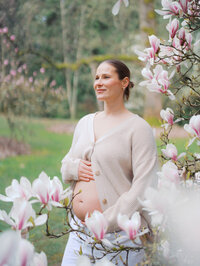 Pregnant woman is standing among blooming magnolias in Seattle, wearing an open knit, showing off her bare pregnant belly