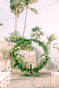 Destination Wedding Photographer captures a floral archway at an outdoor wedding venue with palm trees and string lights in the background.