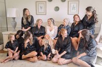 A candid getting-ready moment with the bride and her bridal party at The Sandpiper in Waverly, GA. The bride, wearing a sheer lace robe, sits at the center surrounded by her bridesmaids and flower girls in coordinating black satin robes. Laughter and natural conversation fill the room as they relax together before the ceremony. Captured by Phavy Photography, a St. Simons wedding photographer known for blending editorial detail with candid, documentary storytelling.