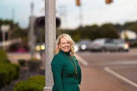 A woman with blonde hair smiles warmly while leaning against a street lamp while standing on a downtown sidewalk. Cars are seen on the road behind her.