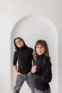 A woman and man stand before a white arched wall, both in black sweaters and jeans. Captured by professional portrait photographers in Washington DC, the woman faces the camera while the man stands slightly behind with hands on his hips.