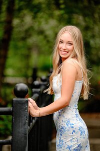 blonde senior girl dressed in a blue and white dress holds onto a bridge railing while looking over her shoulder