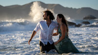 Embracing couple turning to watch the heavy surf approach as the waves crash against the large rocks in the background