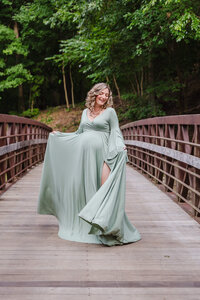 Pregnant woman in a flowing sage green gown standing on a wooden bridge surrounded by trees during a Youngstown maternity photography session