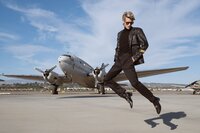 A person wearing a black leather jacket, black pants, and sunglasses is mid-jump in front of a vintage propeller airplane—captured in the bold style of a cinematic portrait photographer Washington DC, under blue skies with wispy clouds.