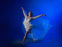 A ballet dancer in a flowing, sheer white dress gracefully extends one leg and raises her arms in a dramatic pose against a vibrant blue background, embodying the elegance of fashion-inspired editorial portraits Washington DC.