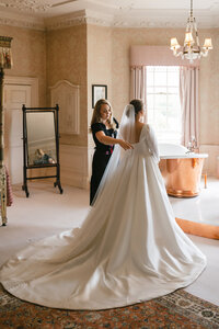 A bride in a white gown with a long train is standing in an elegant room. An attendant helps adjust her dress. The room features a vintage mirror, ornate wallpaper, and a copper bathtub near a window with light pink curtains.