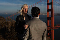 woman facing the camera while the man faces away looking at the golden gate bridge at sunset