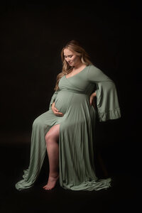 Pregnant woman in a sage green gown standing against a dark studio backdrop during a Cleveland maternity photography session