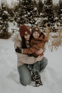 Mom and son laughing together during snowy Maine family photos wearing carhartt hats at a tree farm.