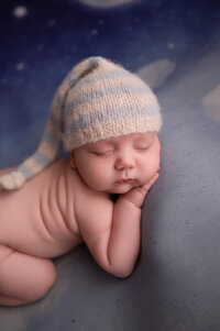 Close-up of a sleepy newborn baby wearing a blue striped knit hat, resting on a soft moonlit background in a professional newborn photography session near Milwaukee, WI.