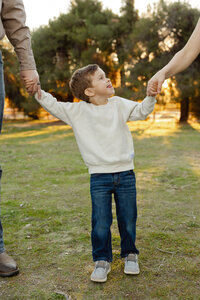 Young boy smiling while holding his parents’ hands in a grassy park at sunset, playful lifestyle family moment photographed by Kaitlyn Dawn Photography.