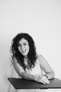 A black and white picture of a woman with dark curly hair smiling brightly as she sits in front of a white backdrop. She wears a white dress and rests her arms on a black table, leaning slightly to her right.