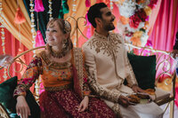 Couple assis côte à côte lors de leur cérémonie du Mehndi, tenues brodées dorées, décor de fleurs colorées et guirlandes en arrière-plan