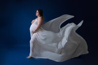 A mother-to-be poses gracefully in a billowing white fabric gown, standing against a deep blue backdrop, exuding strength and beauty.
