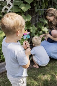 A young boy plays with a pinwheel in the backyard as he looks on his mom and siblings picking vegetables in a backyard garden