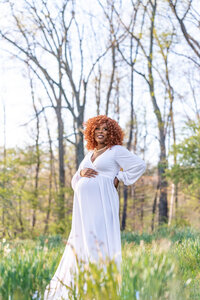 Pregnant woman smiling in a white gown standing in a sunlit wooded field during a Cleveland maternity photography session