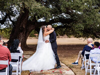 Candid portrait of a bride and groom sharing their first kiss at The Five Oaks Farm in Cleburne during their wedding day by Graceful Ray Photography.