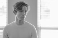 Black and white portrait of a young man in a light sweater standing beside a bright window.