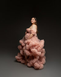 Pregnant woman posing in a flowing blush ruffle maternity gown against a dark background photographed by Kaitlyn Dawn Photography.