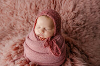 Sleeping newborn baby girl wrapped in a dusty rose swaddle and posed on a soft textured blanket during a studio session with newborn photographer Kaitlyn Dawn Photography.
